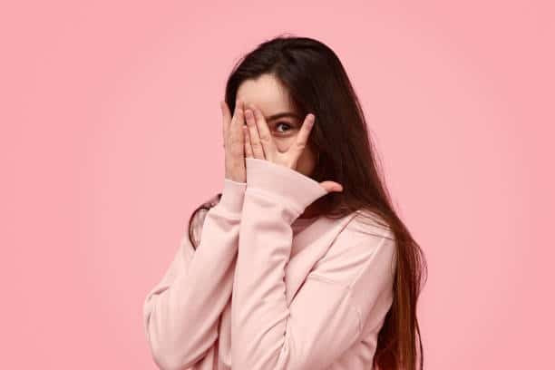 Timid teen girl with long hair covering face with hand and peeking through fingers against pink background